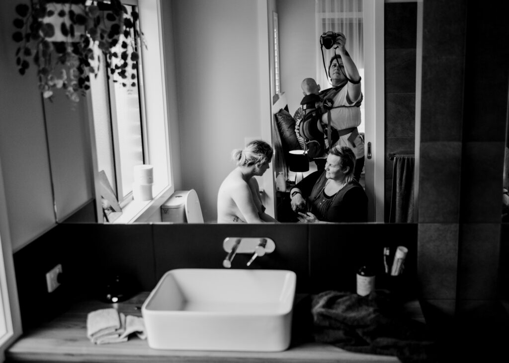 Photo titled 'Working Mums' A midwife and a birth photographer who are a married couple working with a woman at her homebirth in Albury, New South Wales. Their 7 month old baby is being carried in a sling by the photographer. The image is captured in a large mirror's reflection.